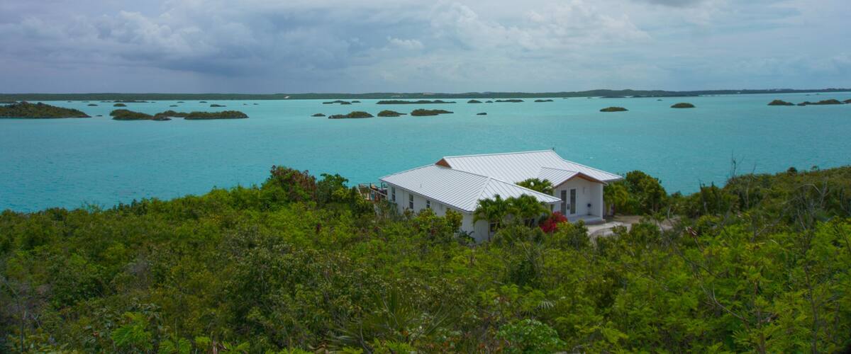 A white house surrounded by lush greenery, overlooking turquoise waters with small islands in the distance.