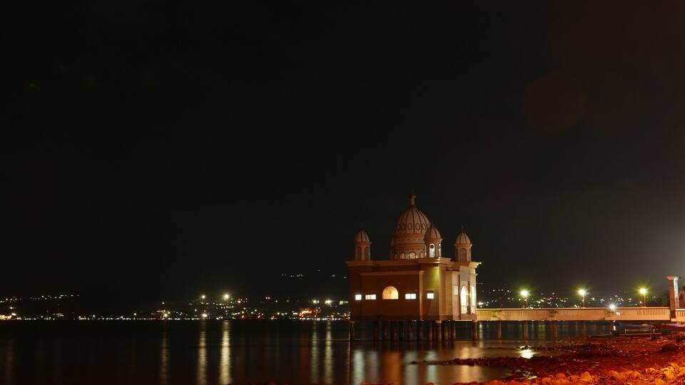 A night view of a domed building with a reflection in the water, surrounded by city lights.