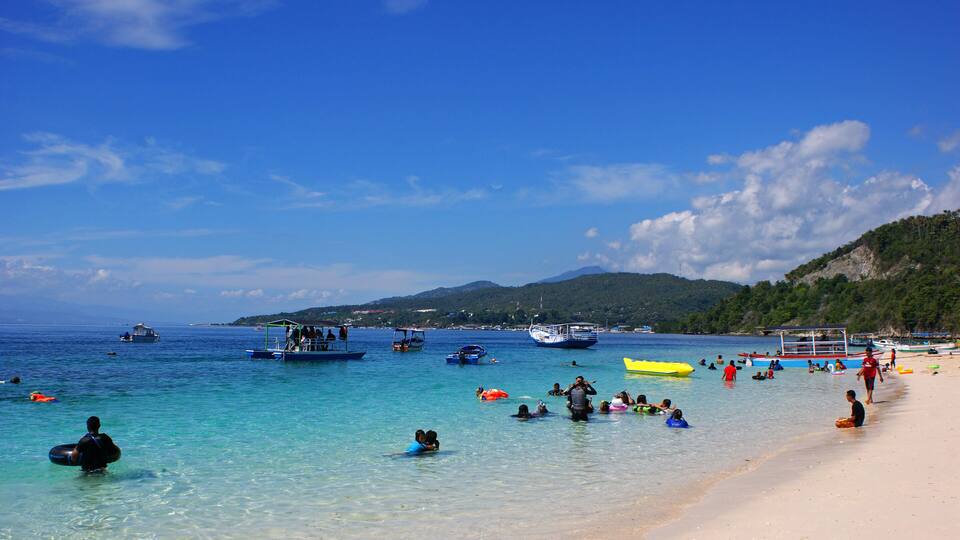 A beach with clear turquoise water, people swimming and sunbathing, and boats in the distance.