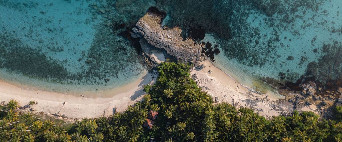 A tropical beach with clear turquoise waters, a rocky outcrop, and dense palm trees.