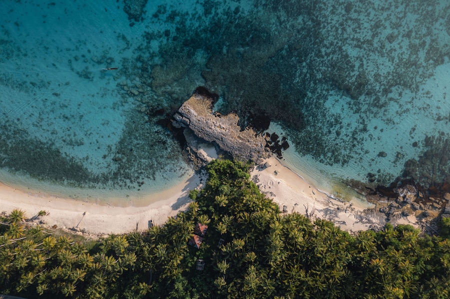 A tropical beach with clear turquoise waters, a rocky outcrop, and dense palm trees.