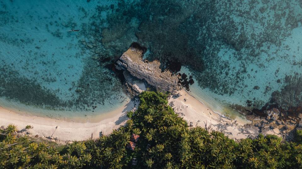 A tropical beach with clear turquoise waters, a rocky outcrop, and dense palm trees.