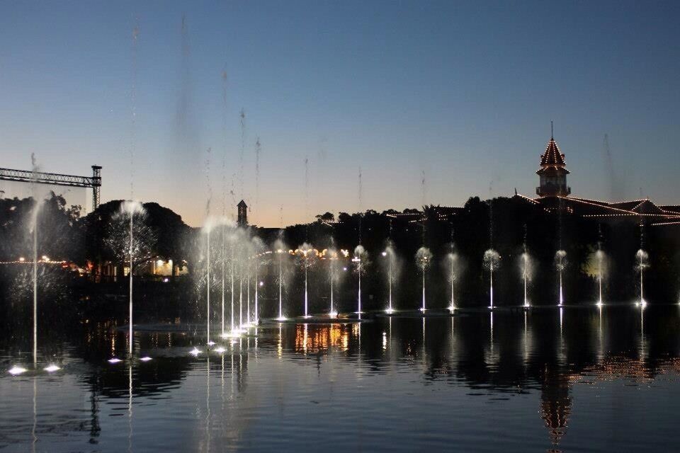Vista nocturna de un espectáculo de fuentes con chorros de agua y un edificio iluminado al fondo.