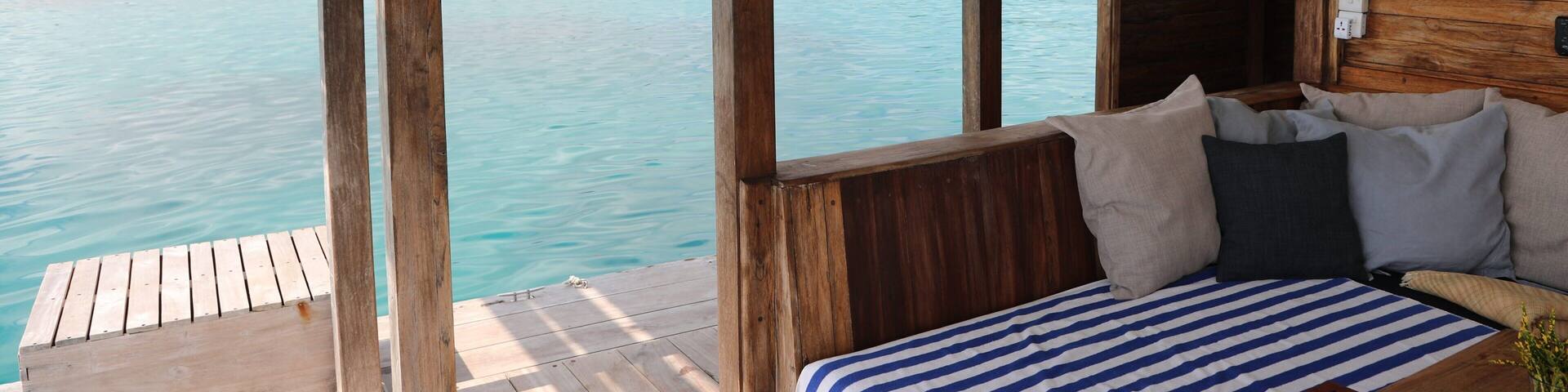 A wooden deck with a bed and a view of turquoise water and a distant shoreline.