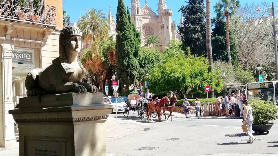 Une statue représentant une figure assise, placée sur une place, avec une cathédrale en arrière-plan et des palmiers tout autour.