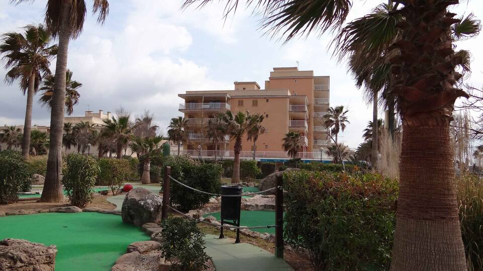 A golf course with a green path, palm trees, and a multi-story building in the background.