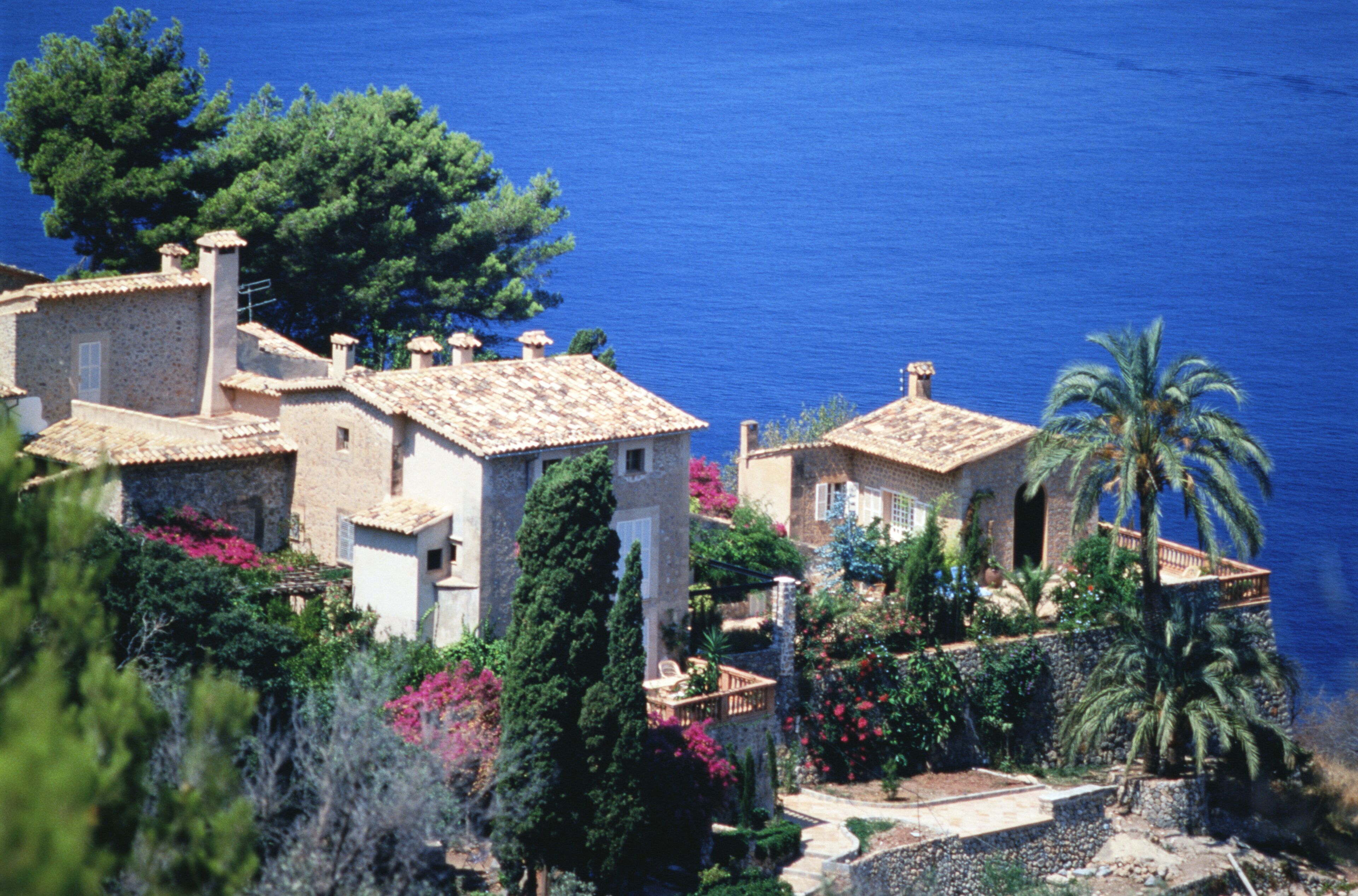 A coastal village with terracotta-roofed houses, lush greenery, and a clear blue sea.