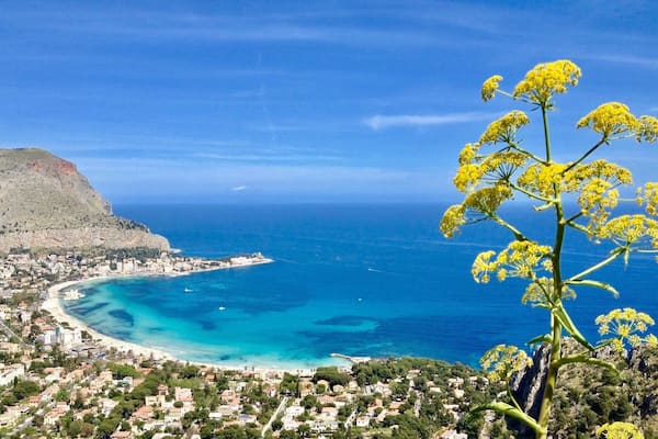 A coastal town with a beach, clear blue sea, and a mountain in the background.