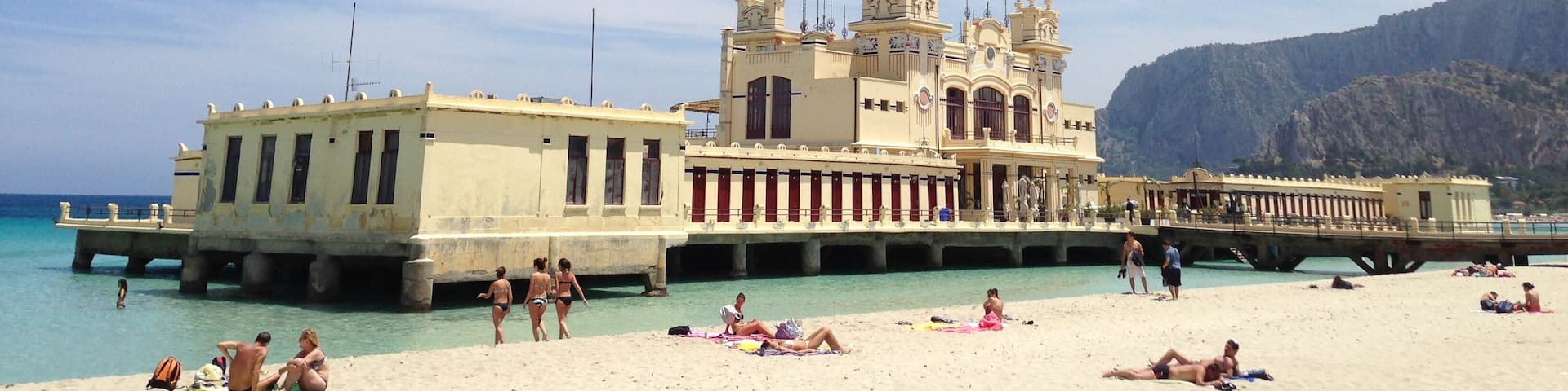A beach with people sunbathing and swimming, a large yellow building with towers, and a rocky mountain in the background.