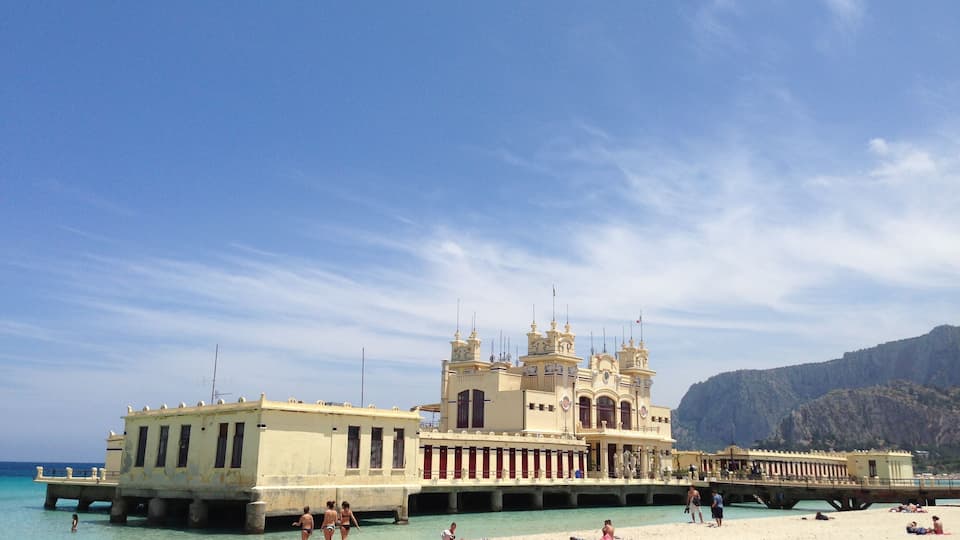 A beach with people sunbathing and swimming, a large yellow building with towers, and a rocky mountain in the background.