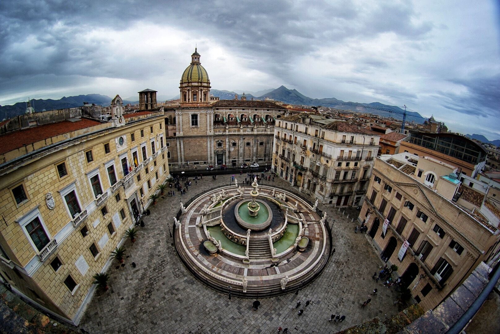 Uma praça histórica com uma fonte central, rodeada por arquitetura clássica e uma montanha ao fundo.