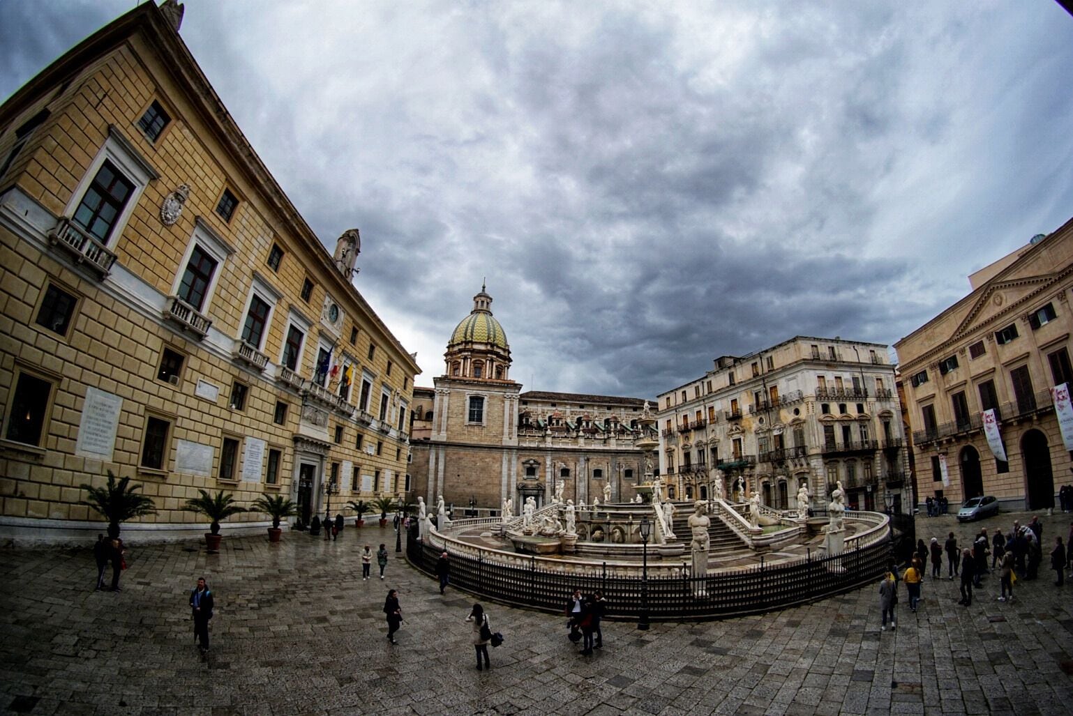 Uma praça histórica com uma fonte, arquitetura clássica e uma cúpula.