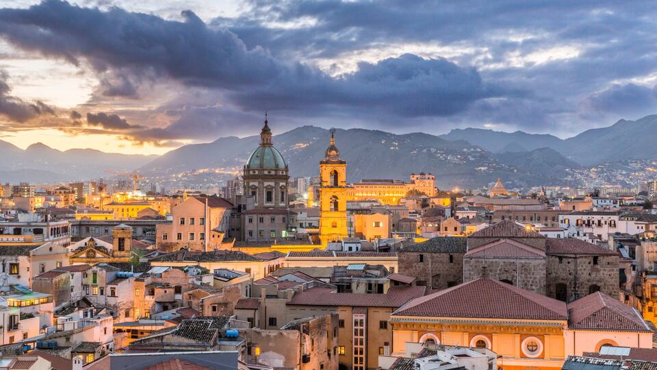 A cityscape with historic buildings, a prominent church with a dome, and mountains in the background.