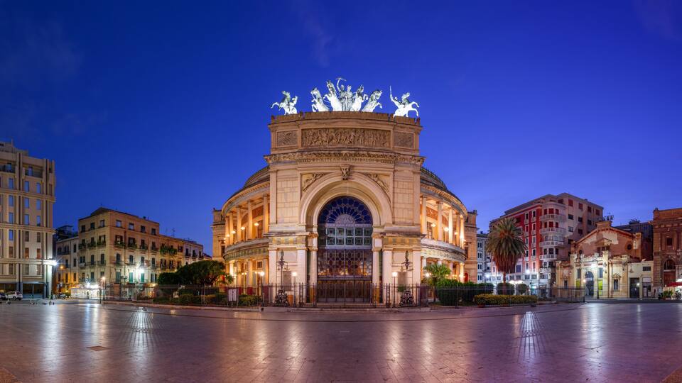 A historic building with a central archway and statues on top, surrounded by other buildings.