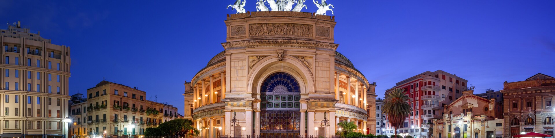 A historic building with a central archway and statues on top, surrounded by other buildings.