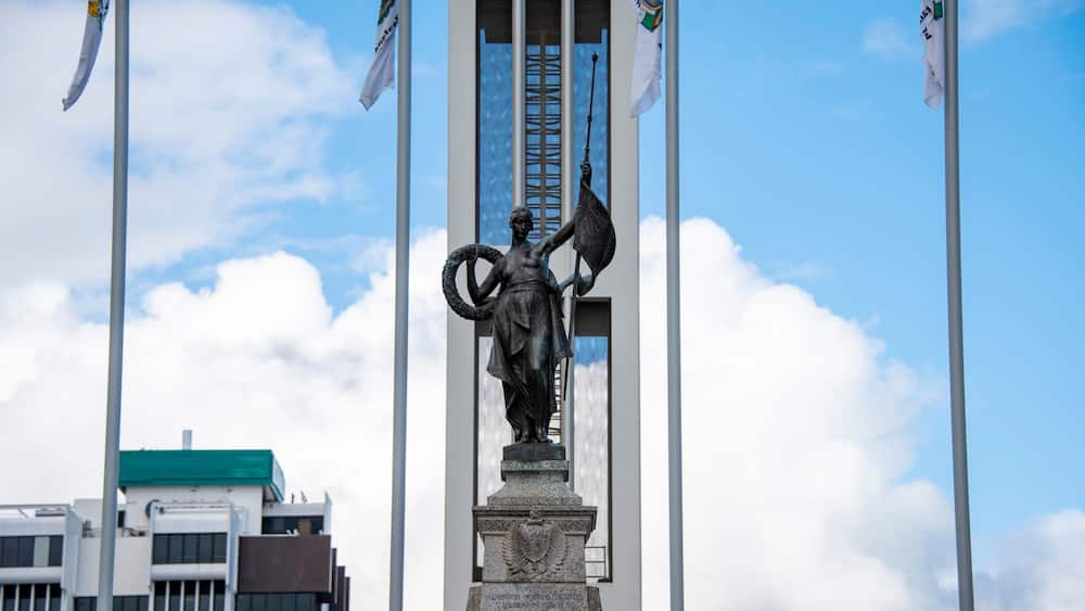 Une statue représentant une figure tenant un drapeau, avec un bâtiment et d’autres drapeaux en arrière-plan.