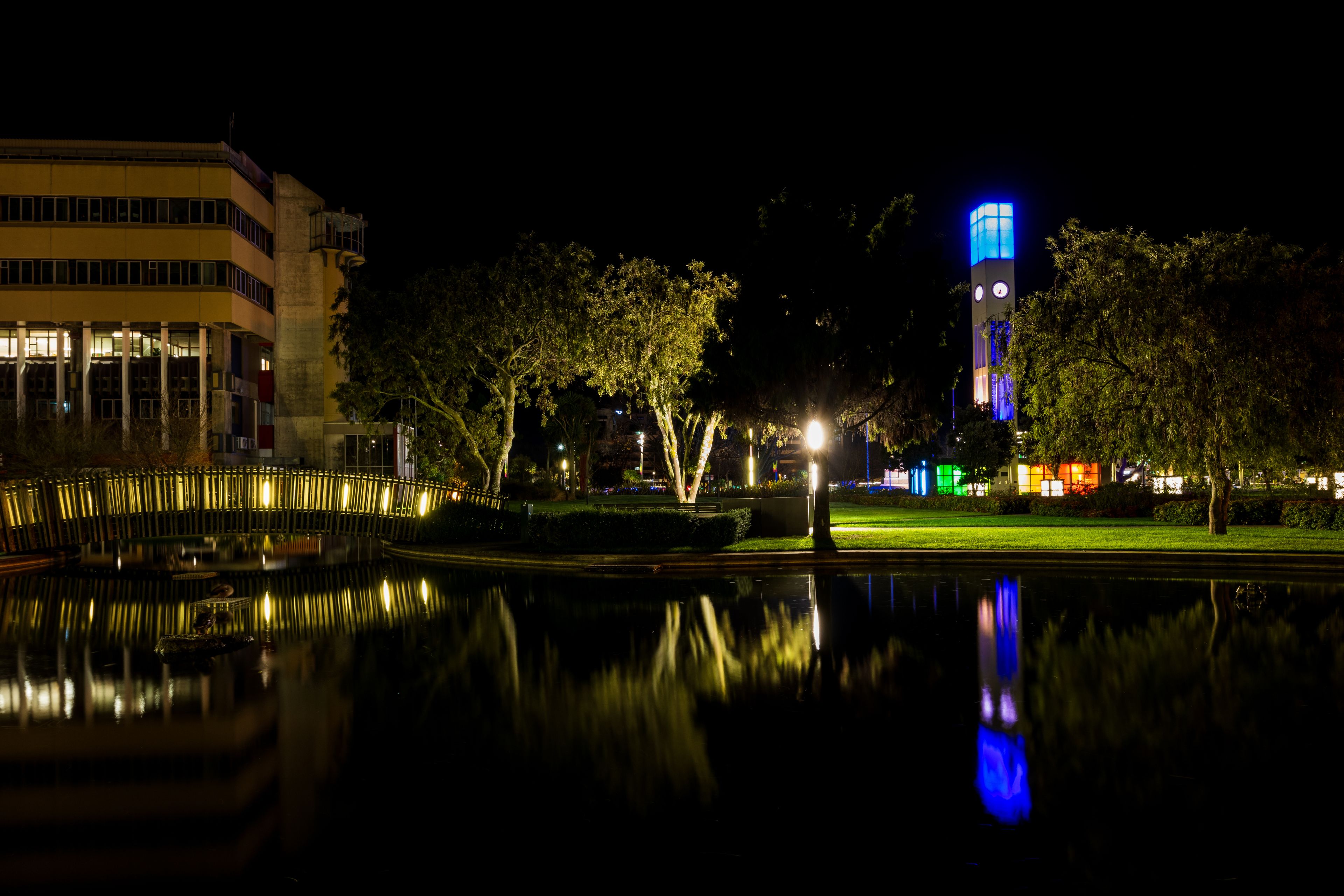 A nighttime cityscape with a brightly lit tower, a calm body of water reflecting the lights, and a building with illuminated windows.