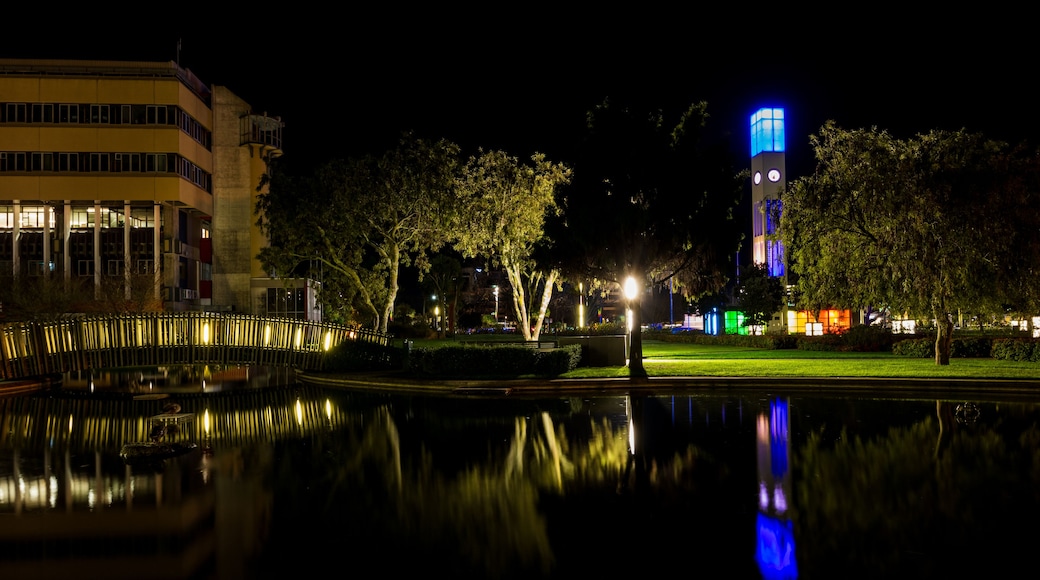 A nighttime cityscape with a brightly lit tower, a calm body of water reflecting the lights, and a building with illuminated windows.