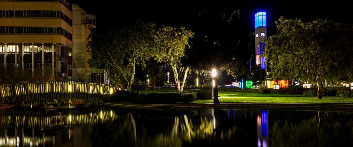 A nighttime cityscape with a brightly lit tower, a calm body of water reflecting the lights, and a building with illuminated windows.