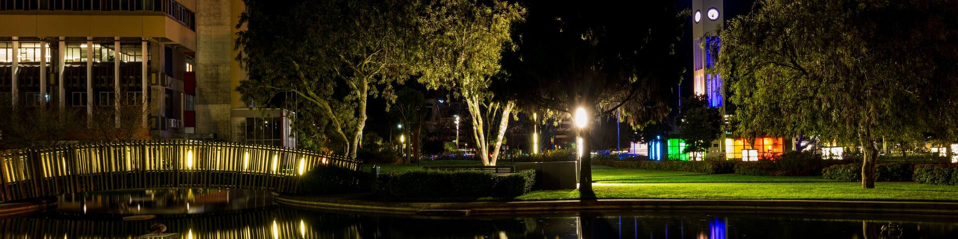 A nighttime cityscape with a brightly lit tower, a calm body of water reflecting the lights, and a building with illuminated windows.