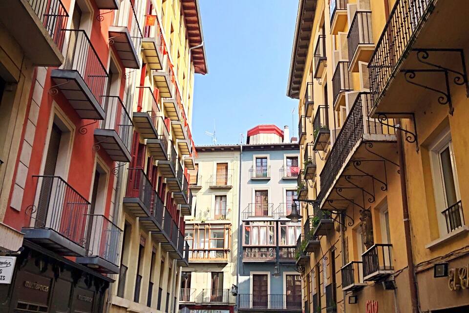 Une rue étroite bordée d’immeubles aux couleurs vives, dotés de balcons, avec une poubelle rouge et quelques piétons.