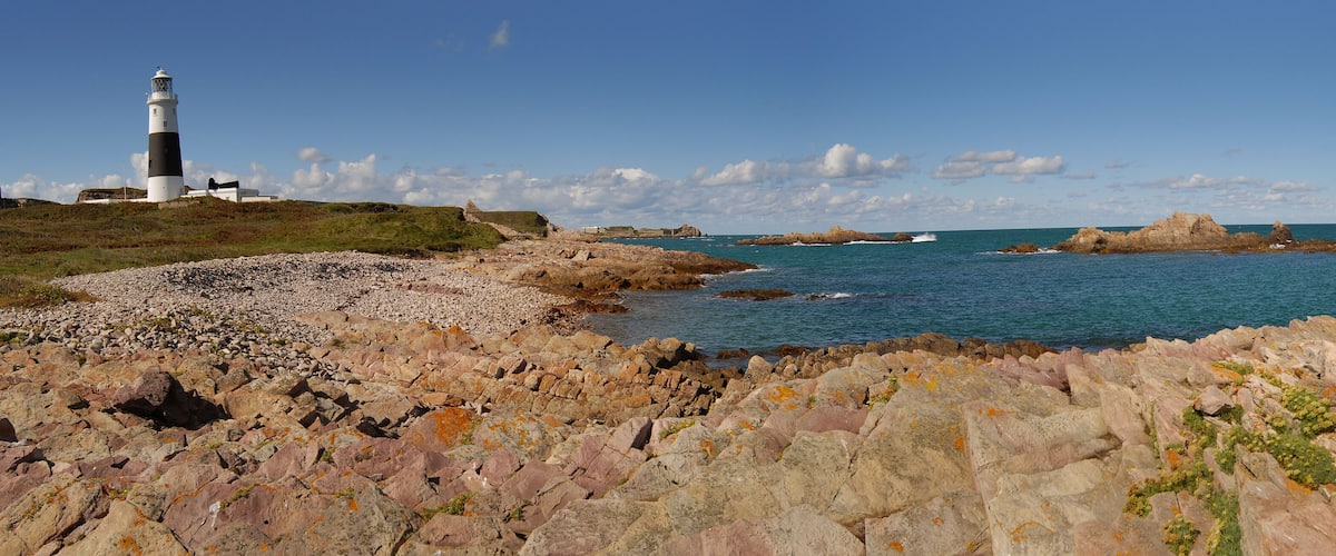 A rocky coastline with a lighthouse, waves crashing against rocks, and a clear blue sky.