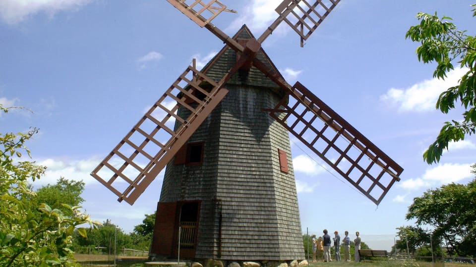 Een traditionele windmolen met houten wieken en een bakstenen basis, omringd door bomen en een heldere lucht.