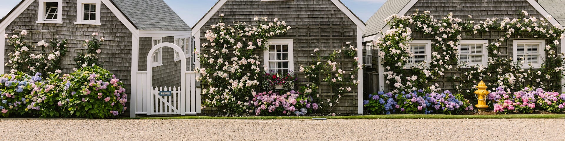 A row of wooden houses with a white picket fence, a flag, and blooming flowers.
