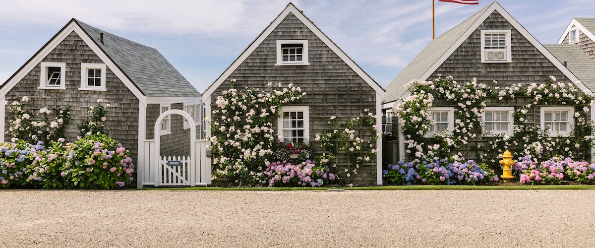 A row of wooden houses with a white picket fence, a flag, and blooming flowers.