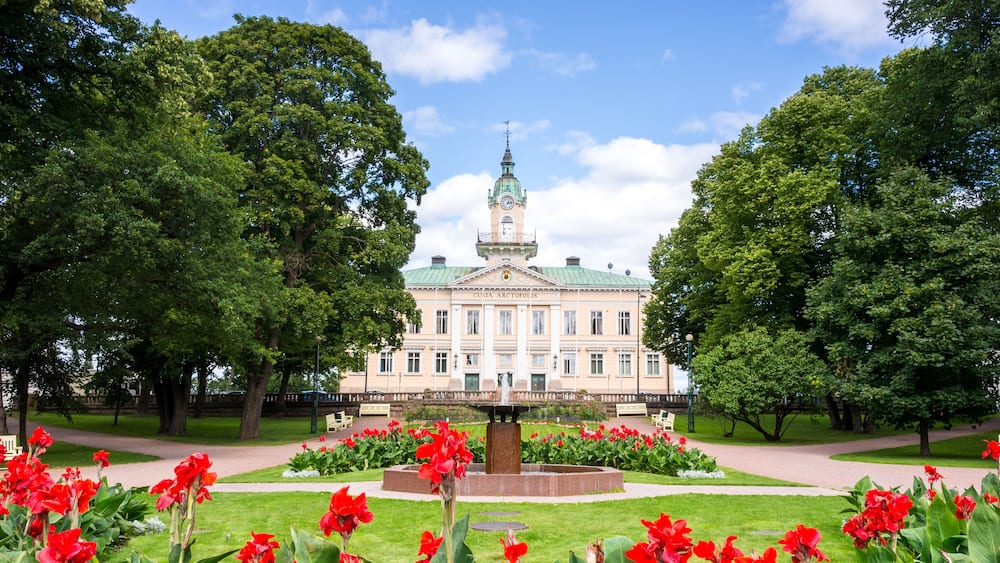 Ein großes Gebäude mit einem begrünten Dach und einem Uhrturm, umgeben von einem gepflegten Garten mit roten Blumen und Bäumen.