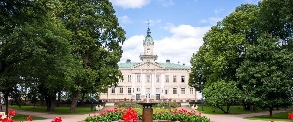 Un grande edificio con tetto verde e una torre dell'orologio, circondato da un giardino curato con fiori rossi e alberi.