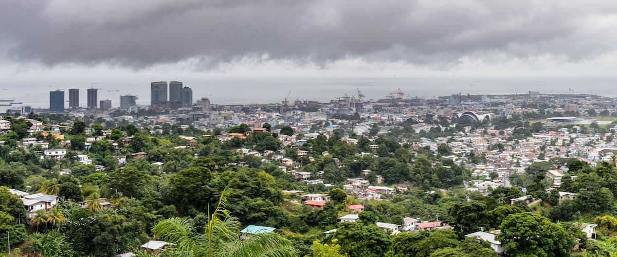 A coastal cityscape with dense housing, a prominent bridge, and high-rise buildings under a cloudy sky.