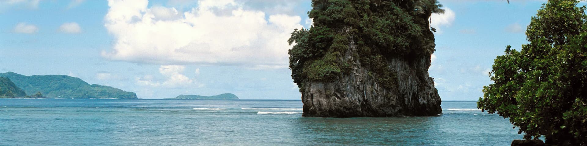 A clear tropical beach with a prominent rock formation surrounded by palm trees and calm sea.