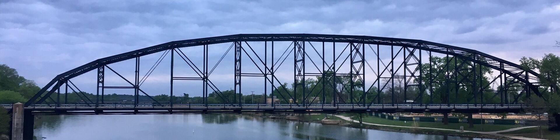 A large steel bridge spanning a calm river with a cloudy sky above.