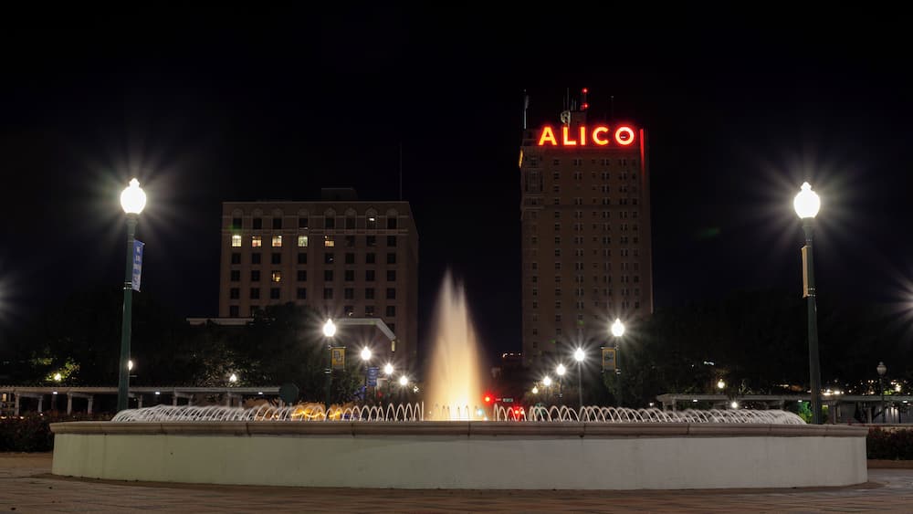 A night scene with a fountain, street lamps, and a building with 'ALICO' illuminated on top.