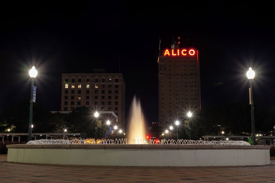 A night scene with a fountain, street lamps, and a building with 'ALICO' illuminated on top.