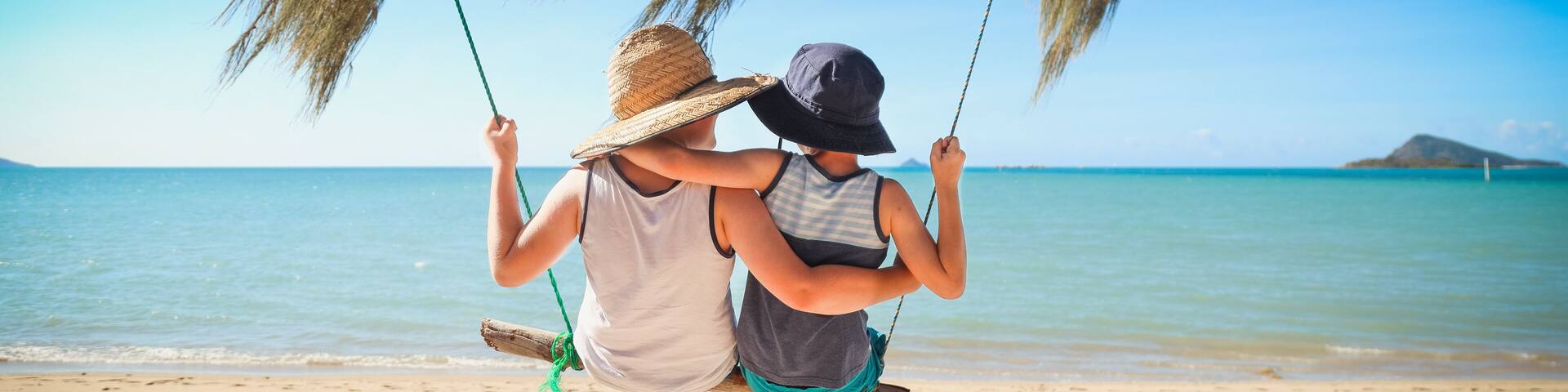 Kids swinging together on log swing at the beach in the Whitsundays, tropical north Queensland. Slow living on tropical family holiday in paradise with sand and sea.