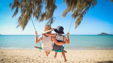 Kids swinging together on log swing at the beach in the Whitsundays, tropical north Queensland. Slow living on tropical family holiday in paradise with sand and sea.