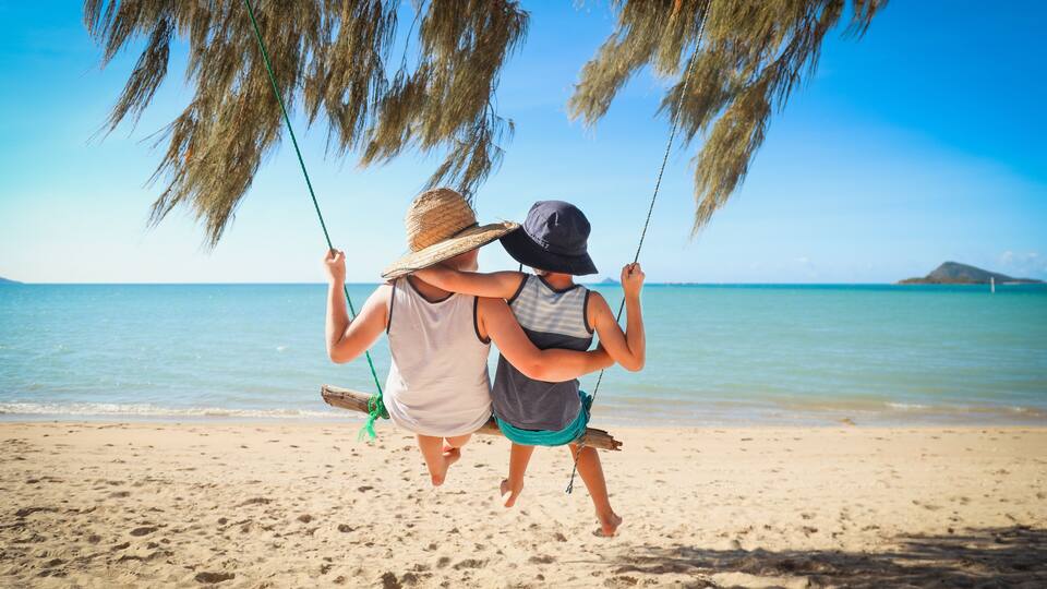 Two people on a beach swing, with a clear blue sky and calm sea in the background.