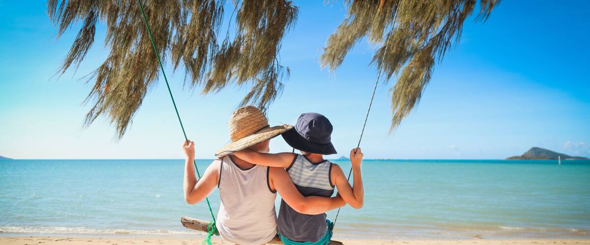 Two people on a beach swing, with a clear blue sky and calm sea in the background.