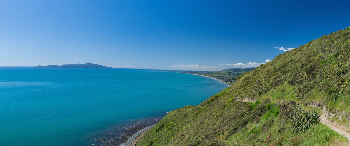 A coastal landscape with a clear blue sea, a winding road, and a green hillside.