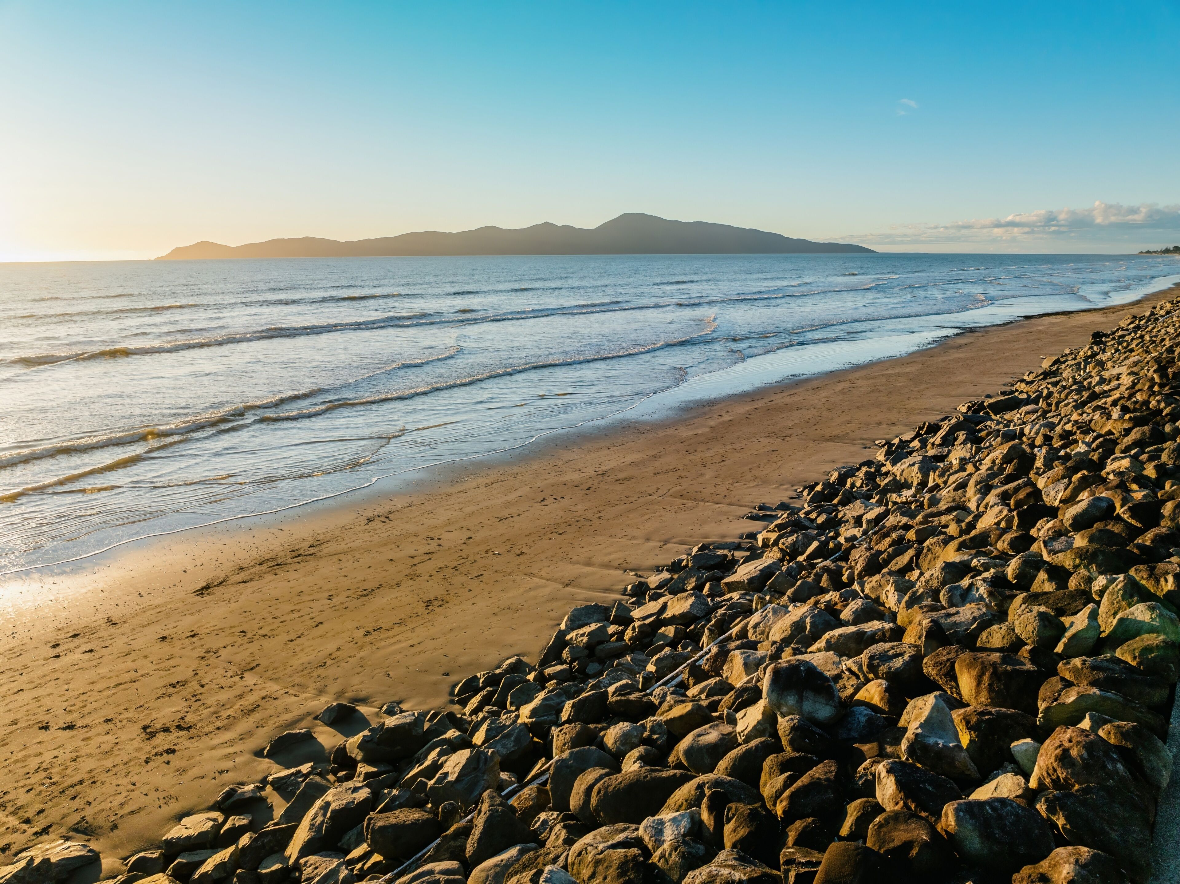 A rocky beach with waves, a clear sky, and distant mountains.