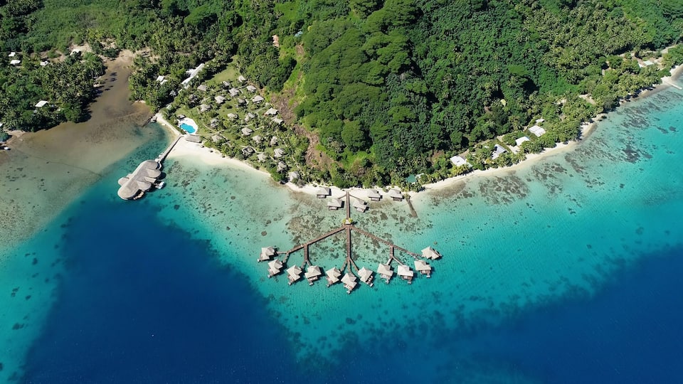 An aerial view of a tropical island with clear turquoise waters, a beach, and overwater bungalows.