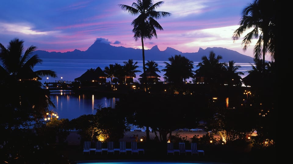A resort with a pool, palm trees, and a view of the ocean at dusk.