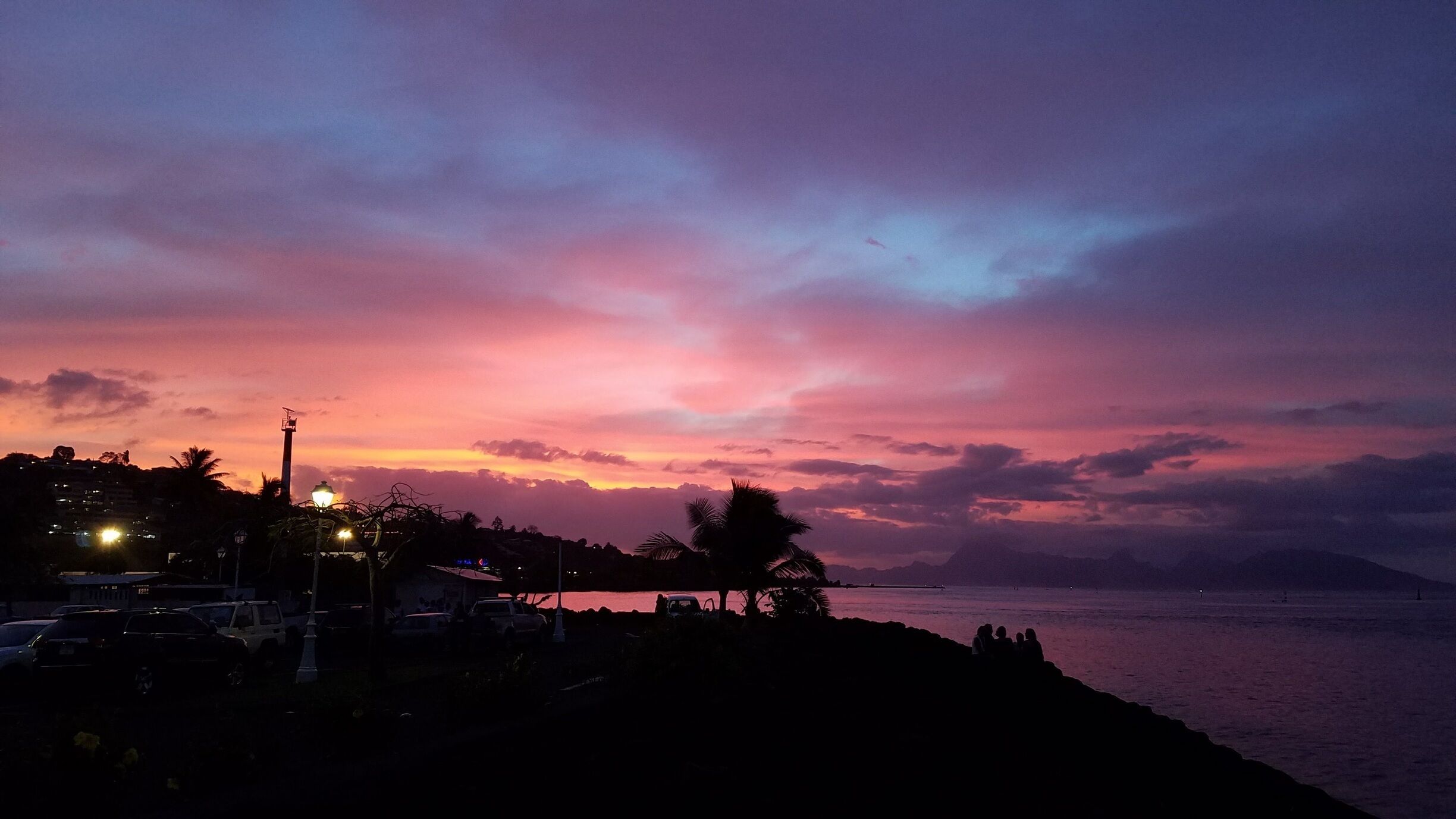A sunset over a coastal area with silhouettes of palm trees, a road with vehicles, and a distant mountain range.