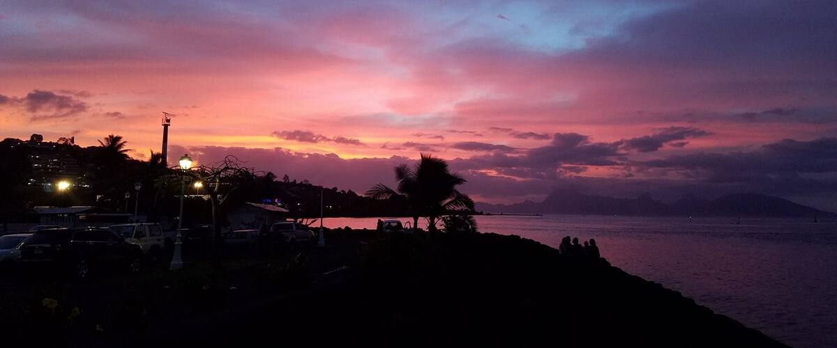 A sunset over a coastal area with silhouettes of palm trees, a road with vehicles, and a distant mountain range.