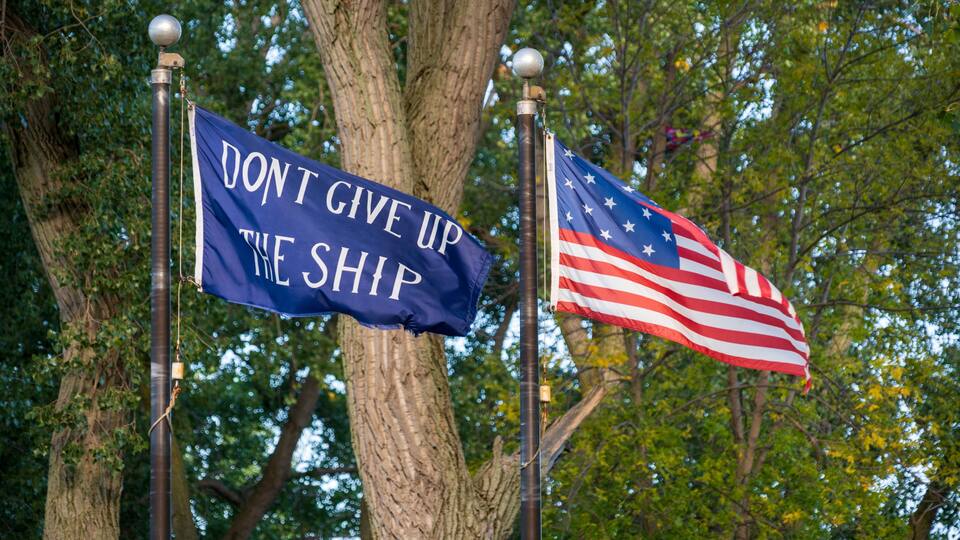 Two flags on flagpoles, one with a blue background and white text, and the other is the American flag.