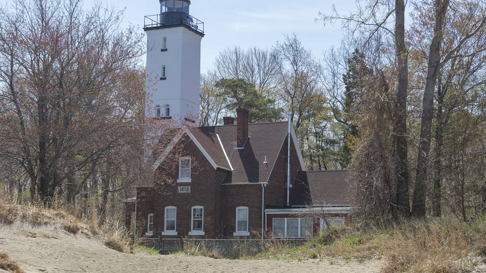 A lighthouse and a brick building with a chimney, surrounded by trees and a dirt path.