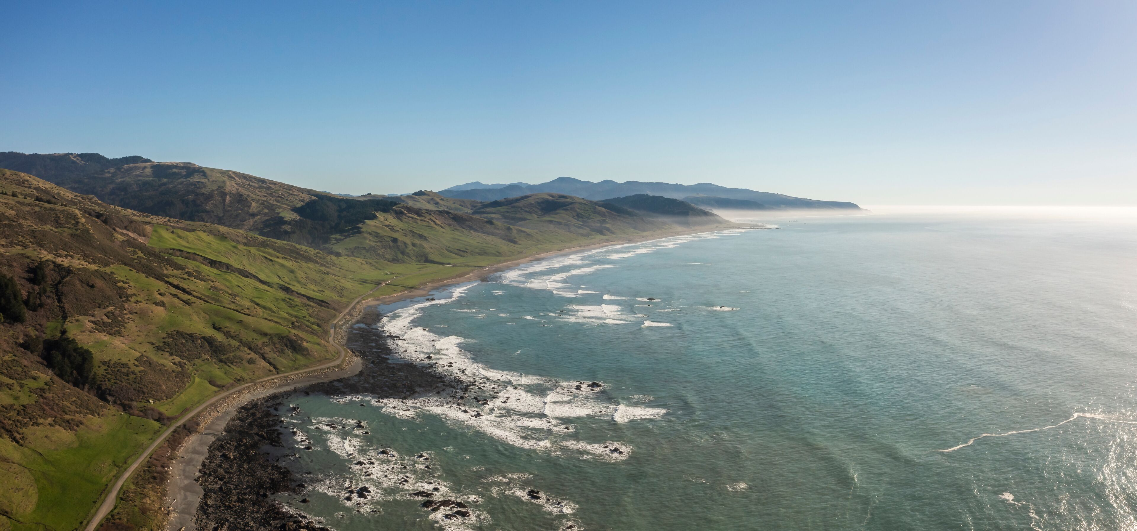 A coastal landscape with a winding road along the shore, rocky outcrops, and distant mountains under a clear sky.