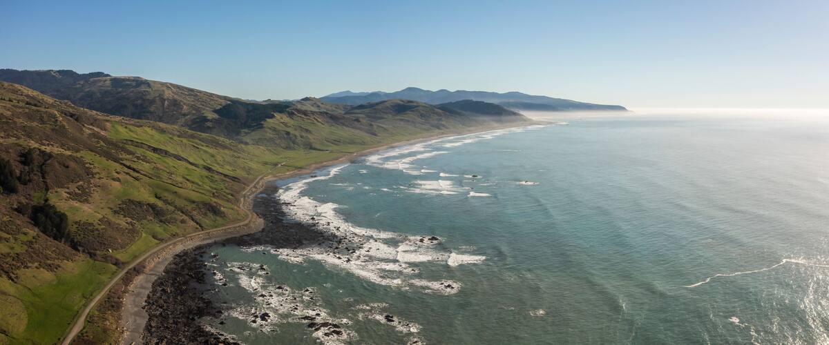 A coastal landscape with a winding road along the shore, rocky outcrops, and distant mountains under a clear sky.